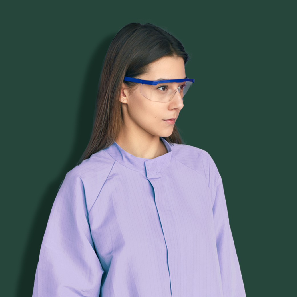 A woman wearing clear safety glasses with a blue frame and a light purple lab coat, shown against a solid dark green background.