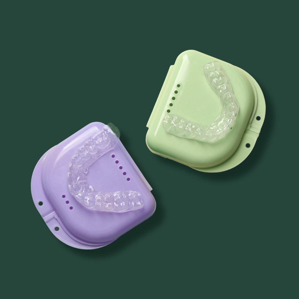 Two clear dental mouth trays resting on matching molded bases, one lavender and one pale green, on a dark green background.